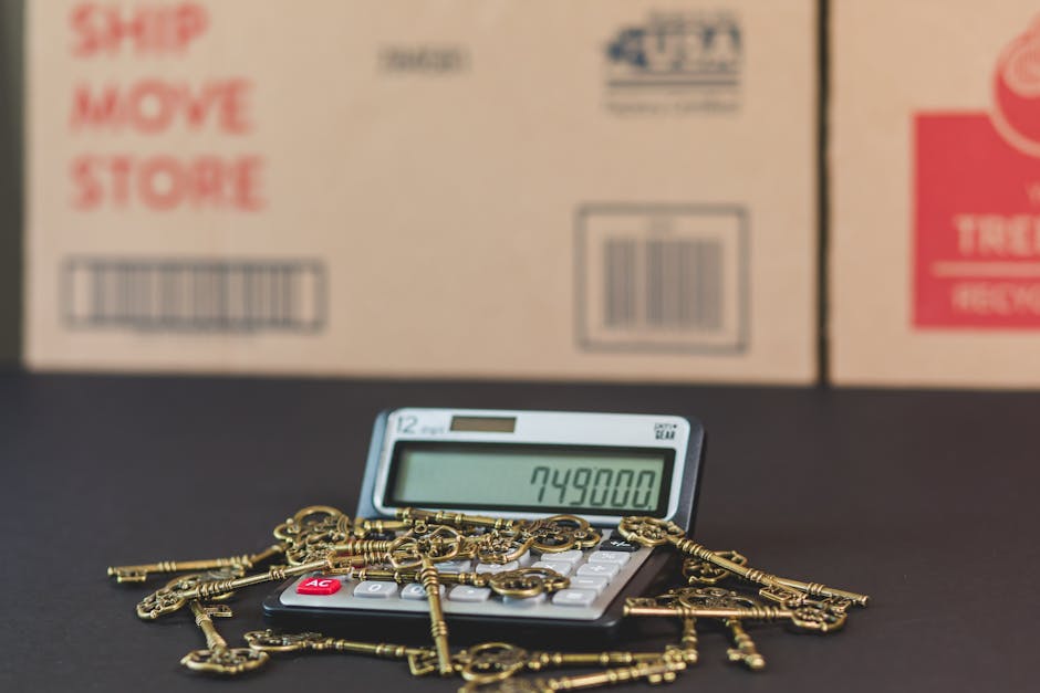 Close-up image of a small electronic calculator displaying the number 749,800, placed on a flat surface. Surrounding the calculator is a scattered pile of small brass keys, some with ornate designs, lying across the calculator and the surface. In the background, there are three cardboard boxes stacked against a wall, each featuring printed labels and barcodes; the boxes are partially out of focus. The scene appears to be indoors, possibly in a storage or packing area, with neutral lighting. This image relates to the process of home relocation and furniture transport, illustrating packing, sorting, or inventory tasks involved in house removals, as provided by Removals Holland Park.