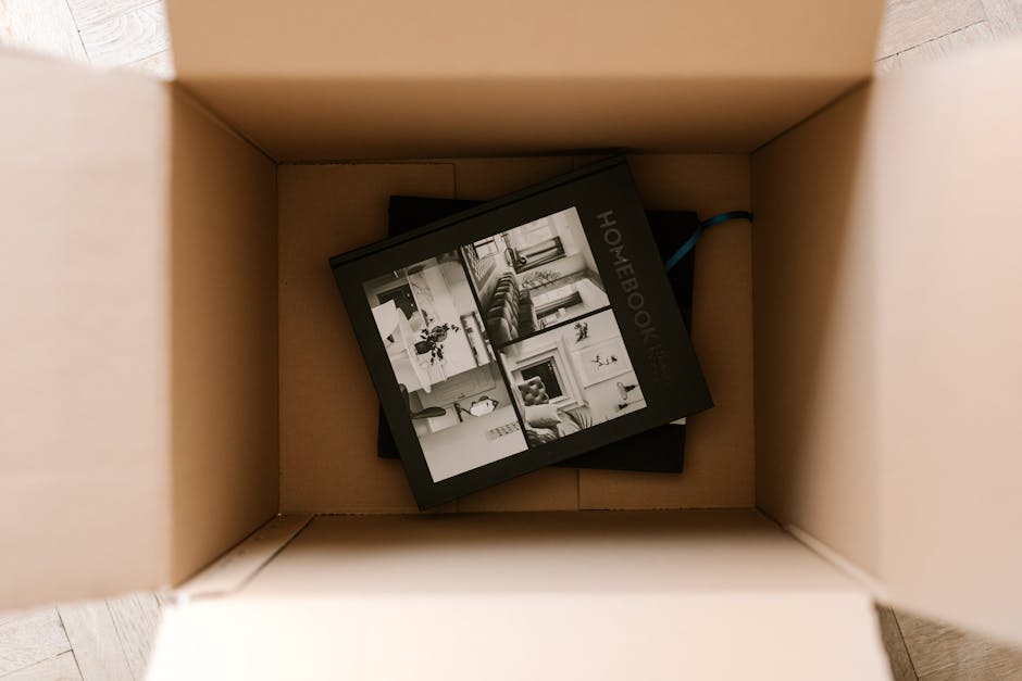 A cardboard moving box viewed from above, with the flaps open inside a home, containing a black and white booklet titled 'Homebook' with images of interior living spaces on its cover. The box is situa