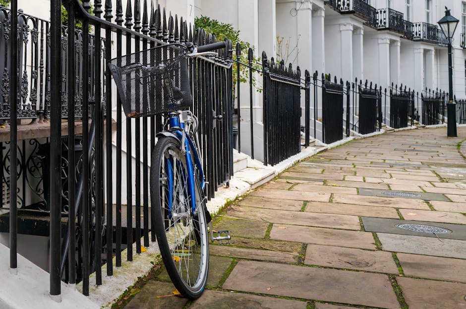 A blue bicycle with a black front basket and silver handlebars is locked to a black metal fence outside a white residential building in a city street. The fence has vertical bars and decorative finials along the top. The pavement consists of large, irregularly shaped stone slabs with some patches of moss and small weeds growing between them. In the background, there are multiple white terraced houses with black iron railings and balconies, some with visible staircases leading up to the entrances. The scene is lit with natural daylight, indicating a clear day. The image reflects a typical urban setting where house removals and packing might encounter obstacles such as narrow pathways, fence barriers, and staircases, as part of the logistics handled by [COMPANY_NAME] during furniture transport and home relocation processes.