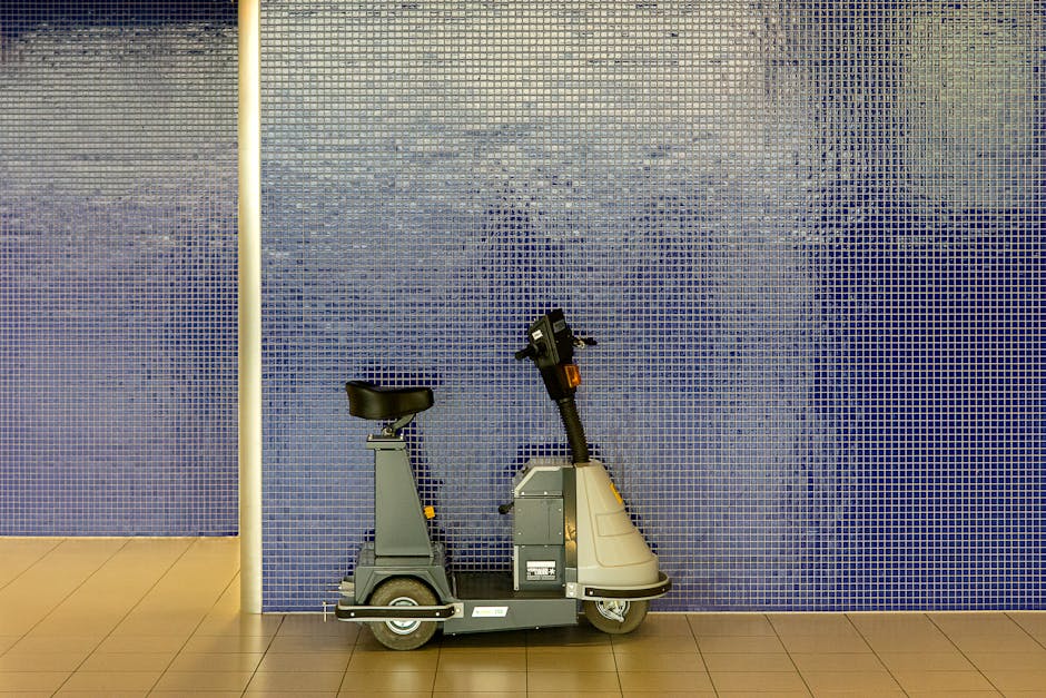 A close-up view of a compact, grey, electric home relocation vehicle parked indoors against a blue mosaic tiled wall. The vehicle features a single seat with a black cushion and a simple control panel at the front handlebar. It is positioned on a beige tiled floor, adjacent to a white vertical support beam, indicating a corridor or lobby space within a residential building. The vehicle is designed for furniture transport and packing and moving services, as utilized by removals companies such as Removals Holland Park. The lighting is bright and evenly distributed, emphasizing the precise details of the vehicle's structure and the textured tiled background, illustrating the logistical considerations involved in house removals and access management during flat moves.