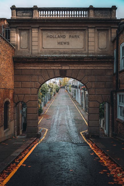 View of the Holland Park Mews entrance with a large stone archway bearing the inscription 'Holland Park Mews' at the top. The cobbled street extends through the arch, flanked by residential houses and narrow pavements on both sides. The street appears to be in the midst of a home relocation process, with visible moving blankets, cardboard boxes, and packed furniture positioned near the doorway of a house on the right. A trolley or hand truck may be seen being used to transport items, and the scene is illuminated by soft natural light, highlighting the preparation and logistics involved in furniture transport along this narrow street. The overall scene reflects careful loading and packing activities by Removals Holland Park, fitting within the context of a professional house removal or moving service.