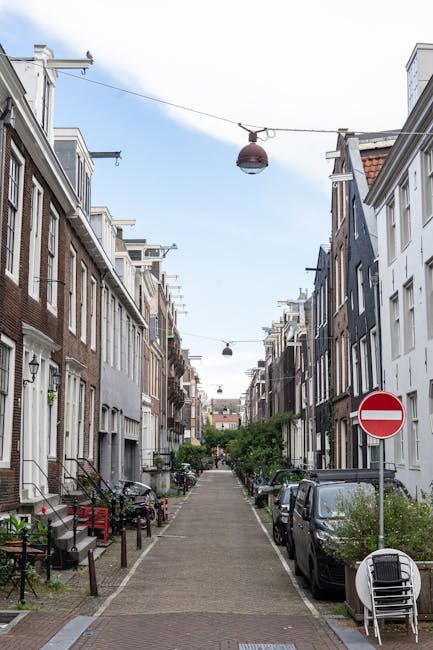 A narrow residential street on Holland Park Road, with multi-storey brick buildings on both sides. The pavement is made of interlocking brick, and bicycles are parked along the sidewalks. Some greenery, including potted plants and small trees, is visible on the building ledges and storefronts. A sign reading 'Sozan' is attached to one of the buildings on the right. Several pedestrians are seen walking along the street, with a few near the entrance of a property and others further down the road. The sky above is partly cloudy, and the street is well-lit with natural daylight. This setting depicts an urban environment suitable for house removals and furniture transport, with visible outdoor packaging materials and the initial stages of a home relocation process, supported by services such as those offered by Removals Holland Park.