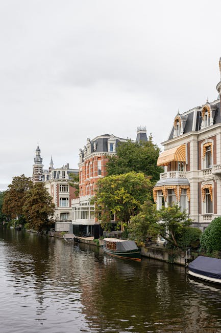 The image shows a row of historic residential buildings along the canal in Holland Park, with architectural details including ornate facades, bay windows, and decorative roof structures. The buildings are made of brick and stone, with some featuring striped awnings and large windows. The canal in the foreground reflects the buildings and the cloudy sky above, while a narrowboat and a small private boat are moored close to the water's edge. Trees with green foliage line the canal, partially obscuring lower parts of the buildings. The scene is well-lit by natural daylight, capturing the peaceful setting typical of London’s house and apartment exteriors. This setting exemplifies the type of property involved in house removals, with furniture, packing materials, and moving equipment typically used by removals Holland Park services visible during the home relocation process.