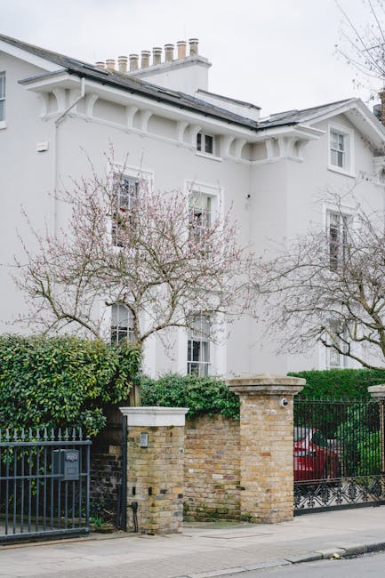 A street view in Holland Park showing a row of residential buildings behind a brick and wrought iron fence, with flowering trees in full bloom along the pavement. The buildings are three stories high, with large windows and decorative architectural details. A yellow bus stop sign is positioned on the sidewalk near the fence. The scene is illuminated by bright daylight, with shadows cast by the trees and street lamps. This exterior environment depicts a quiet neighbourhood suitable for house removals, as [COMPANY_NAME], specialising in removals and home relocation services, might coordinate the logistics of furniture transport and packing within this area. No moving equipment or activity is visible in the image, focusing instead on the residential setting that could be involved in a house move or packing process, consistent with the tips provided for Ilchester Place house removals in Holland Park.