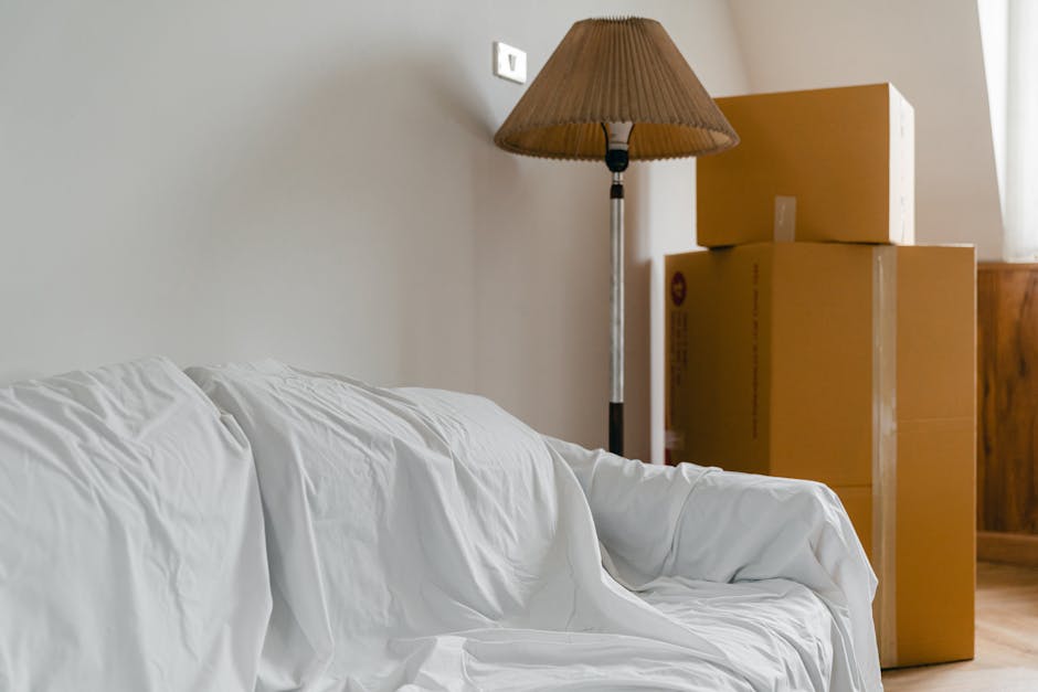 Indoor scene showing a white sofa covered with a protective slipcover positioned against a plain light-colored wall. To the right of the sofa, there is a tall standing lamp with a brown pleated lampsh