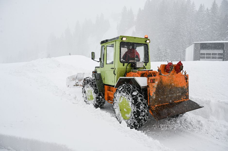 A green and orange snowplough vehicle with large snow tires is clearing a thick layer of snow from a driveway or street, with snow-covered ground and trees in the misty, foggy background. The vehicle has a front-mounted orange plough blade actively pushing snow aside, and the cabin has dark-tinted windows. In the background, a modern grey garage or storage building is partially visible, located in a residential or commercial property area. The scene is illuminated by natural light under overcast skies, with snow falling and accumulating on the vehicle and surroundings. This image captures the process of snow removal, typical during winter weather conditions, which may relate to services offered by providers such as Removals Holland Park when responding to urgent or immediate needs for clearing pathways or access routes during house relocations or emergencies involving heavy snowfall.