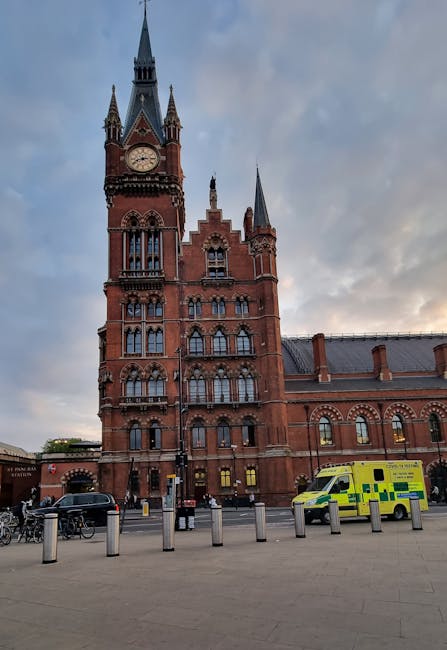 A large red-brick Victorian-style building with a prominent clock tower featuring pointed spires, set against a cloudy sky during late afternoon or early evening. In front of the building, on a paved pavement, several cardboard boxes and wrapped furniture items are visible, suggesting packing preparations or an ongoing home relocation process. A yellow and green emergency vehicle is parked nearby, alongside a few other cars and bicycles, with short bollards separating the street from the pedestrian area. The scene appears to be part of a relocation or moving operation, with no visible personnel or loading equipment present, and the environment is well-lit with natural daylight. The image captures the urban setting associated with moving services, with the historic architecture complementing the scene of packing and transport activities overseen by a professional removals company like Removals Holland Park.
