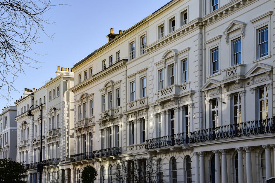 A row of elegant white Victorian-style terraced houses with ornate architectural details, including decorative window frames, small balconies with black wrought-iron railings, and classical mouldings. The buildings have large sash windows and are situated along a paved street under a clear blue sky. A black street lamp is visible on the pavement in front of the buildings. The scene appears to be during daytime with natural sunlight illuminating the facades. The image captures the exterior of properties in Holland Park, with a focus on architectural features relevant for house removals or furniture transport. The context is suitable for discussing packing, loading, and home relocation processes that Removals Holland Park may provide for flats or houses in this area, highlighting the structural environment for moving services.