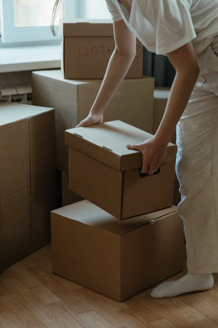 A person in light clothing, with their arms extended, is seen lifting a cardboard box labeled 'CLOTHES' from a stack of similar boxes inside a well-lit room near a window with light curtains. The boxes are plain brown, sealed, and stacked on top of one another on a wooden floor. Some boxes appear to contain household items, likely prepared for a home relocation or moving process. The individual is wearing white socks and is engaged in the packing and moving activity, with additional boxes visible nearby. This scene illustrates the packing stage of a house removal, supported by the professional services offered by Removals Holland Park, part of their removals and furniture transport solutions tailored for flats in areas like Holland Park.